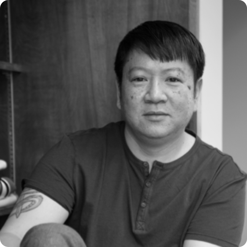 Black and white portrait of a person with short hair and a tattoo on their upper arm, wearing a short-sleeved, buttoned shirt, sitting in front of a wooden shelf.