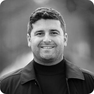 A man with short dark hair smiles at the camera, wearing a dark jacket over a turtleneck. The background is blurred in grayscale.