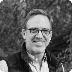 A man with short hair and glasses, wearing a collared shirt and a vest, smiles outdoors in front of a blurred leafy background. The image is in black and white.