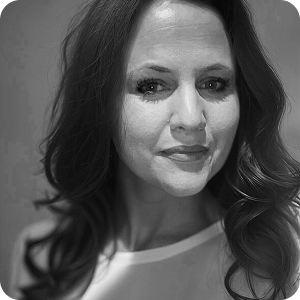 Black and white portrait of a woman with long, wavy hair, wearing a light-colored top, looking at the camera with a slight smile. The background is blurred.