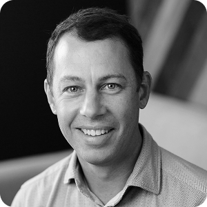 Black and white portrait of a man with short hair, wearing a collared shirt, smiling at the camera with a blurred background.