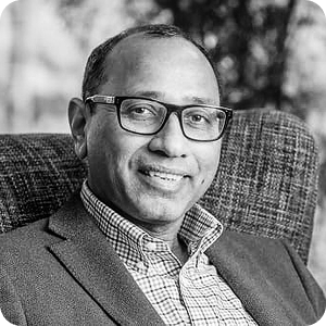 Black-and-white photo of a man wearing glasses, a checked shirt, and a blazer, smiling while seated in a textured armchair. Trees and outdoor scenery are blurred in the background.