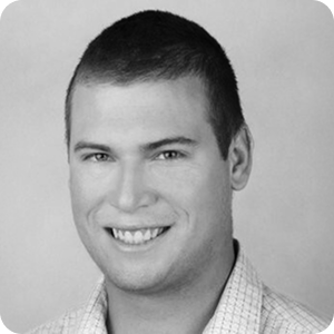 A young man with short hair smiles at the camera. He is wearing a light-colored, checkered shirt and the background is plain and light. The image is in black and white.