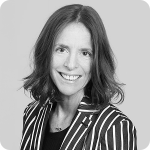 A woman with shoulder-length brown hair smiles at the camera. She is wearing a black and white striped blazer over a dark top, against a plain light-grey background.