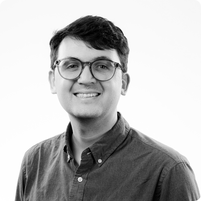 A young adult with short dark hair, wearing round glasses and a button-up shirt, smiles at the camera against a plain white background.
