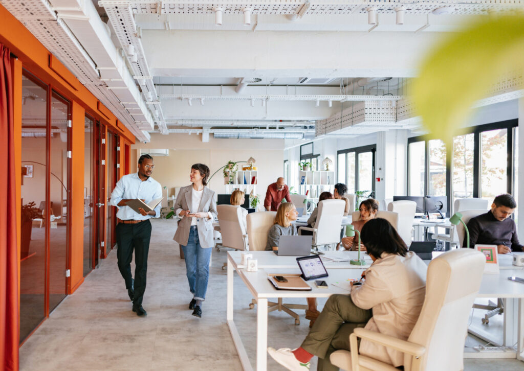 Modern open office space with several people working at desks and collaborating; two colleagues walk and talk, while others focus on laptops or paperwork. Large windows provide natural light, and plants decorate the space.