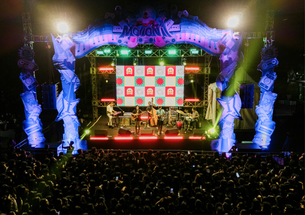 A band performs on a brightly lit stage with colorful decorations and a large screen displaying graphic patterns, as a large crowd watches at an outdoor night concert.
