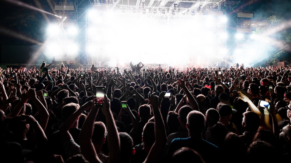 A large crowd at a concert with many people holding up phones, silhouetted against bright stage lights and smoke, creating an energetic and lively atmosphere.