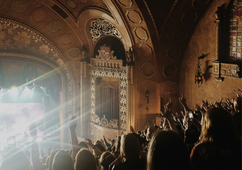 A crowd inside an ornately decorated theater cheers and raises their hands toward a brightly lit stage, with beams of light streaming across the venue’s arched ceiling and carved accents.