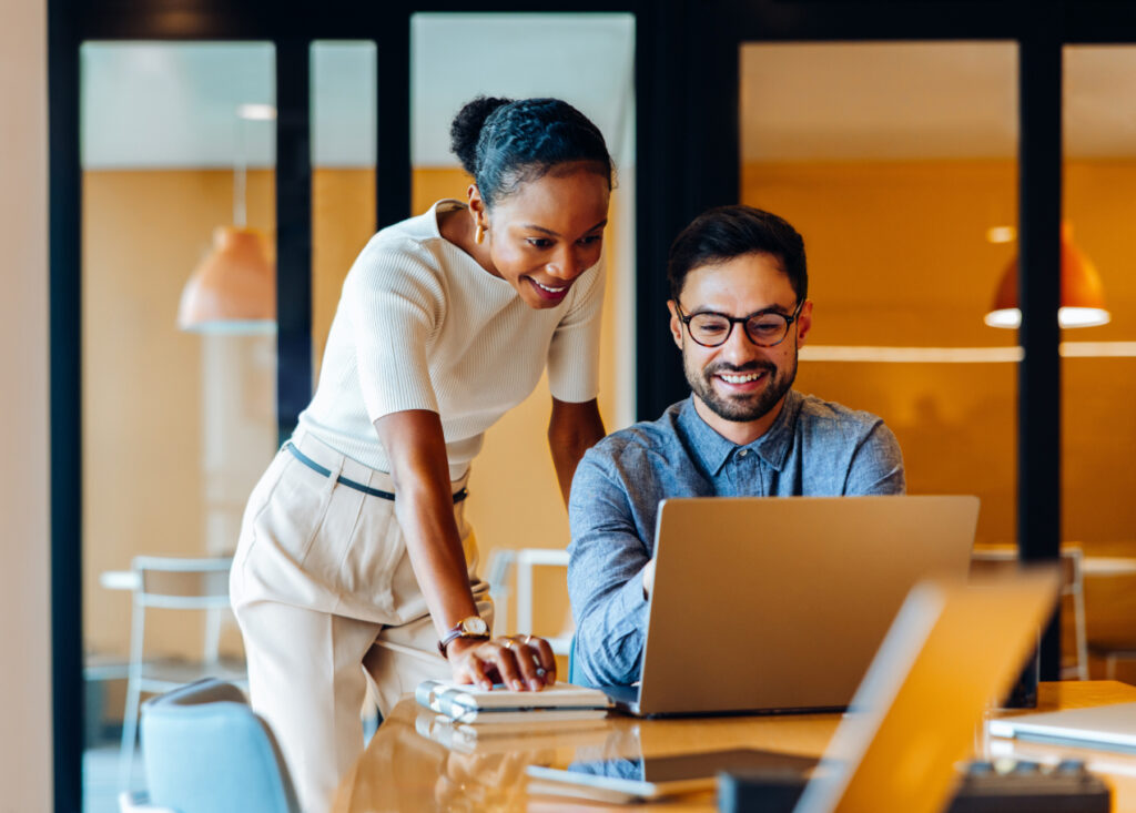 Two colleagues in an office, one standing and one sitting at a table, look at a laptop screen and smile. The workspace has glass walls and warm lighting in the background.