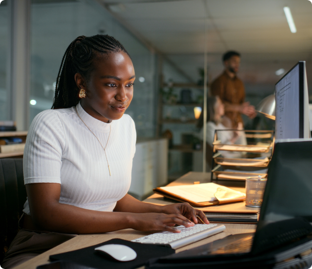 A woman with braided hair sits at a desk in an office, typing on a keyboard and looking at a computer monitor. Papers and office supplies are on the desk, and two people are visible in the background.