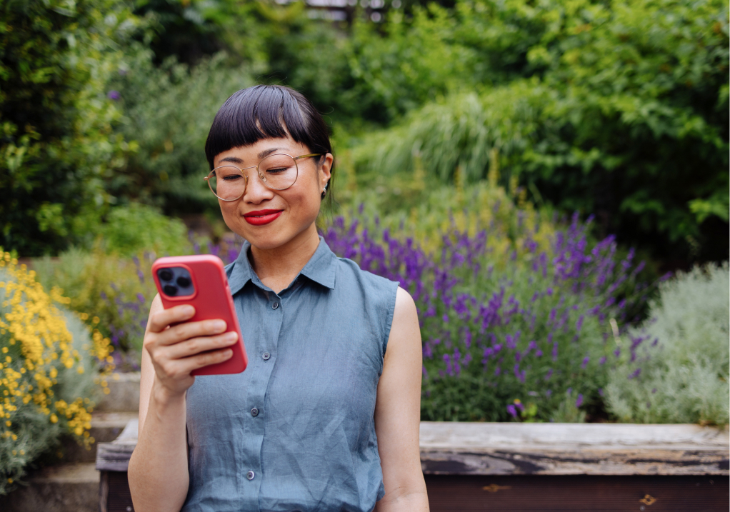 A woman with short dark hair and glasses smiles while looking at her smartphone in a garden with purple and yellow flowers and lush greenery in the background.