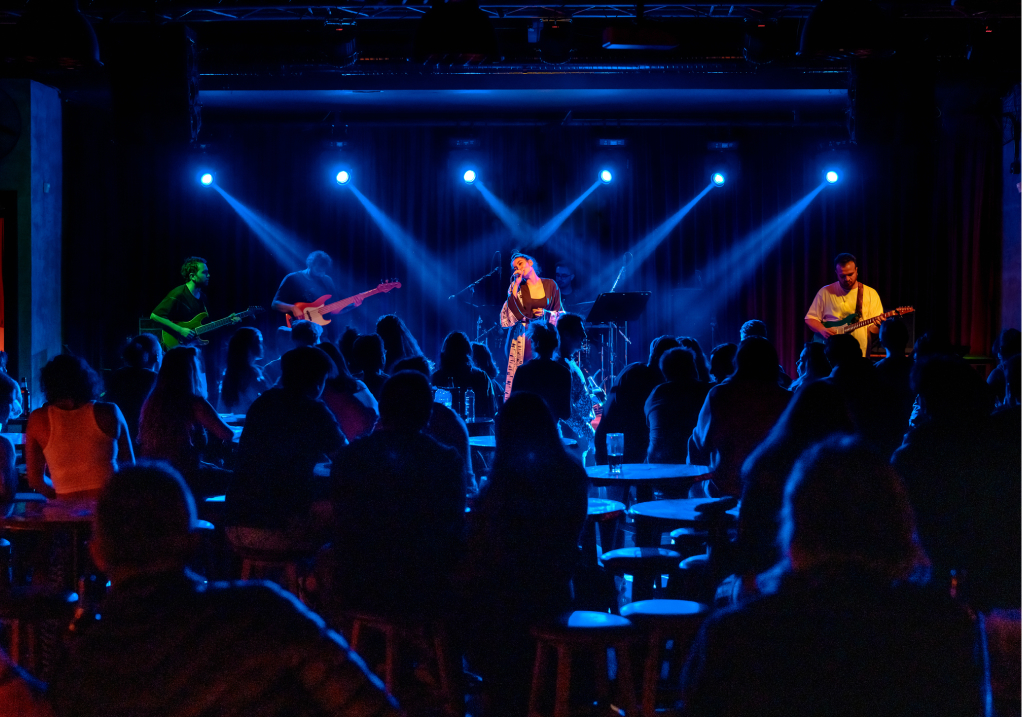 A band performs onstage under blue lights in front of an audience seated at tables in a dimly lit music venue. The scene captures the lively atmosphere of a live concert.