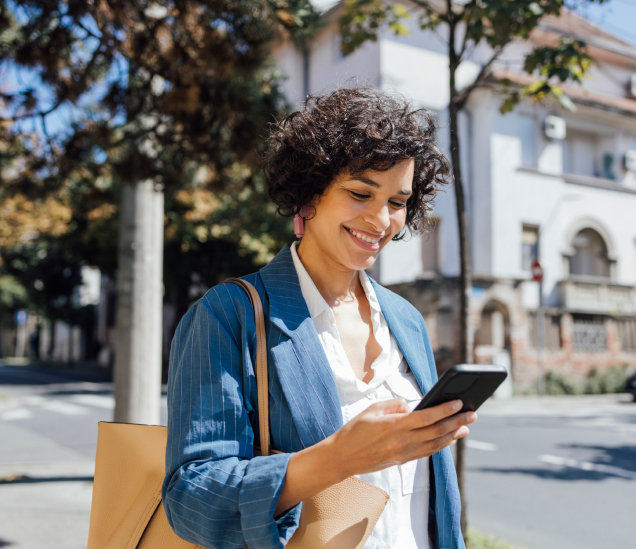 A woman with curly hair, wearing a blue blazer and carrying a beige bag, smiles while looking at her smartphone outdoors on a sunny day, with buildings and trees in the background.