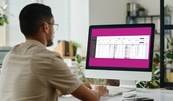 A man with glasses sits at a desk using a desktop computer, viewing a spreadsheet or data table on the screen in an office setting with plants and shelves in the background.