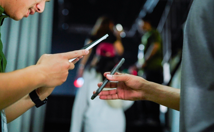 Two people stand close together, holding and using smartphones. Their faces are partially visible, and the background is blurred with two more people visible further away. The setting appears casual and indoors.