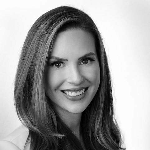 Black and white portrait of a woman with long, straight hair, smiling and looking directly at the camera. She is wearing a light-colored top and the background is plain and bright.