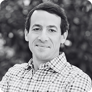 A man with dark hair, wearing a checkered shirt, smiles softly at the camera. The background is blurred with what appears to be trees or foliage. The image is in black and white.